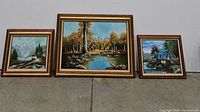 Three framed nature-themed oil paintings displayed side-by-side, showing mountain, cabin, and cottage scenes.