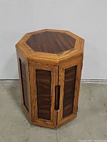 Frontal view of octagonal side table showing darker wood top and side panels framed by lighter wood. Doors are closed revealing cut-out handles on sides.