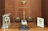 Photo showing brass weighing scale, Bulova anniversary clock, Lenox porcelain clock, and metal cannon figurine arranged on wooden shelf