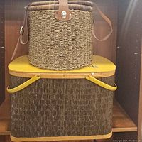 Two baskets stacked in shelving. Bottom is rectangular Burlington Basket Company vintage picnic basket with yellow lid and handles. Top is smaller round wicker basket with leather strap.