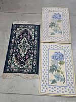 Three small rugs laid out on floor; one blue-green floral rug with fringe and two cream rugs with blue hydrangea floral design and blue polka dot border.