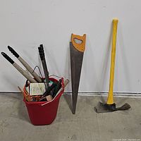 Full view of red bucket with several hand tools, handsaw with orange handle, and ax with yellow handle and black head