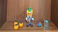 Overall lot photo showing the clown figurine, three glass oranges, paperweight, and keepsake all displayed on a wooden shelf.