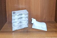 Two packages of white ceramic cupcake pedestal plates stacked, next to a white ceramic cow butter dish on a wood shelf.