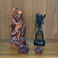 Four carved wood figurines displayed on a wooden surface against a wooden backdrop. From left to right: tall Asian elder with staff and bird, black abstract human figurine, small reddish elephant, and laughing Buddha figurine.