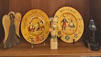 Shelf display showing wooden angel, two hand painted folk art plates, and a wooden figurine of a woodsman.