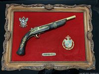 Full view of framed imitation pistol with all items on red fabric background inside carved wooden frame