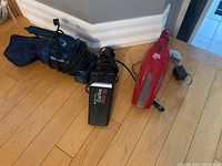 Three vacuums on wooden floor: red Dirt Devil handheld vacuum, black vacuum with cloth bag, and CarVac vacuum with attached cords