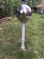 Outdoor backyard statue with large reflective metal sphere on ornate pedestal standing on grassy lawn with garden background.