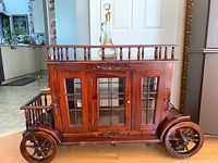Front view of wooden bar cart showing carved front panel, glass-paneled double doors and decorative wagon wheels.
