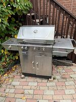 Outdoor older stainless steel BBQ grill with lid closed, thermometer on lid, three control knobs, two side burners with control knobs on sides, and cabinet storage below. Shows signs of outdoor use.
