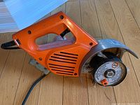 Orange Black and Decker handheld grinder lying on wooden floor, showing side profile with ventilation slats and power cord.