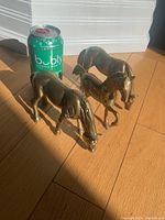Three brass horse figurines shown alongside a green sparkling water can for size comparison. Horses are positioned naturally with detailed sculpting visible.