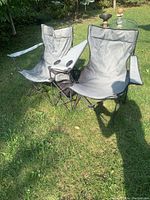 Front view of two connected camping chairs side-by-side on grass, showing gray fabric and black frame with center drink and storage holder.