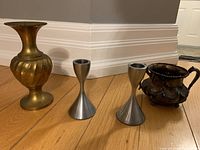 Group photo of one brass vase, two silver metal candle holders, and one small decorative metal cup with handle, placed on wooden floor with white baseboard background.