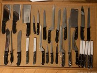 Wide view of all knives laid flat on a surface showing full lot content of 29 pieces of assorted cutting tools primarily knives and cleavers.