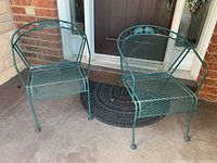 Two green metal chairs placed outdoors on a concrete surface in front of a door, showing mesh seat and back design with curved armrests.