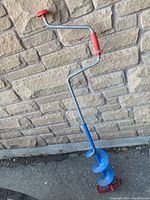 Photo showing full manual ice fishing auger drill with red handle and blue cutting blade against a stone wall background.