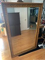 Full view of large ornate gold-framed mirror standing on floor reflecting room interior.
