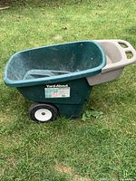 Side view of a green plastic wheelbarrow with gray handles and two wheels. The Yard-About label is visible on the side of the tub. The wheelbarrow is placed outdoors on grass, showing functional but light use signs.