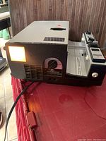 Front-left angled view of the vintage black Sawyers Rotomatic 747AQ slide projector showing illuminated lamp and front lens on a red table with brown wooden panel background.