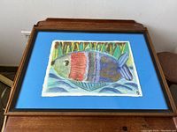 Framed abstract fish painting by Hanni Rothschild with blue mat and brown wooden frame placed on wooden surface.