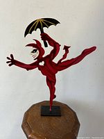 Front and side view of red enameled devil figure sculpture holding umbrella, standing on octagonal wooden stool.