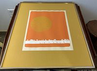 Framed orange and gold serigraph print with a yellow mat and silver frame, placed on wooden furniture against a white wall.