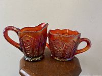 Pair of iridescent red purple glass items - cream jug and sugar bowl - displayed on wooden surface