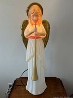 Front view of vintage blow-mould angel figure playing trumpet, illuminated from inside, white gown with pink sash, gold wings and halo.