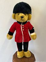 Full front view of the 18 inch vintage Merrythought teddy bear dressed in red Buckingham Palace Guardsman uniform jacket and tall black fur hat, standing on a wooden surface.