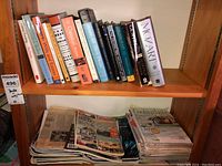 Shelf with a variety of books and a stack of magazines beneath. Books include music topics and literature, magazines appear to be farming and cooking focused.