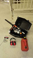 Overall photo showing the Echo CS-3450 chainsaw inside a black plastic carrying case, red 3M ear mufflers, and a red gas/oil mix can on a concrete floor.