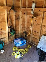 Wide view of corner showing Kobalt cordless blower in box, two string trimmers (Arnold and Ryobi), yellow rope and yellow measuring tape on wooden floor surface