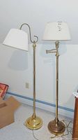 Two brass finish floor lamps, one with a straight post and tapered shade, the other with a curved arm and pleated shade.