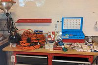 Wide view of workbench with orange extension cords, black clamp lights, blue multimeter case and various electrical connectors and parts spread on table.