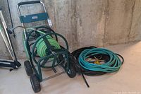 Left side view of green metal hose cart with garden hose spool and adjacent black coiled hoses