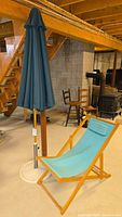 Full view of vintage wooden folding lounge chair with blue canvas fabric and accompanying large blue patio umbrella with heavy white metal stand, placed indoors on concrete floor.
