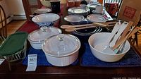 Overall view of lot on table showing blue patterned Pyrex nesting bowls, white Corningware baking dishes with lids, Revere Ware mixing bowls, wooden spoons, and whisks on a blue cloth.