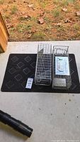 Two Safeguard metal wire animal trap cages and one black rubber doormat placed on concrete with grass background.