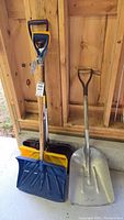Four snow shovels propped against wooden wall, visible from above mid-handle down to blade, included are blue and yellow plastic shovels and a metal shovel with wooden handle.