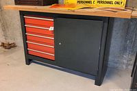 Front view of the tool cabinet with five red drawers and black lockable cabinet door beneath the plywood top.
