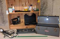 Craftsman toolbox, handheld scroll saw, socket wrench case, and workshop light with cords on wooden shelf and concrete floor