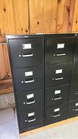 Front view of two black metal vertical file cabinets with 4 drawers each, showing label holders and pull handles, placed against wood wall.
