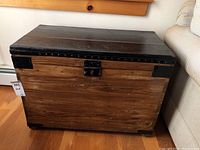 Front view of the vintage wooden chest showing wood grain and black top with metal latch and corner reinforcements.