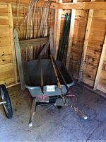 View of wheelbarrow with three garden tools in tub, tomato plant cages and fencing in background inside wooden shed.