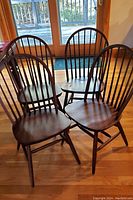 Four wooden Windsor chairs with spindle backs arranged on hardwood floor in front of glass doors allowing natural light.