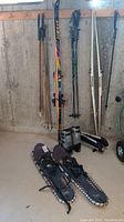 Full view of skis, poles, snowshoes, and boots leaning against basement wall.