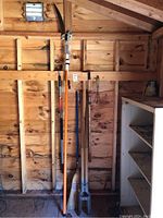 Wide shot showing six yard tools hung on a wooden wall inside a shed including a pole pruner, broom, two hoes, and a post hole digger.