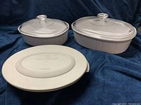 Three Corningware casserole dishes: two with glass lids (medium and large oval shapes), one with white plastic lid (small round shape). All white ceramic with ribbed sides.